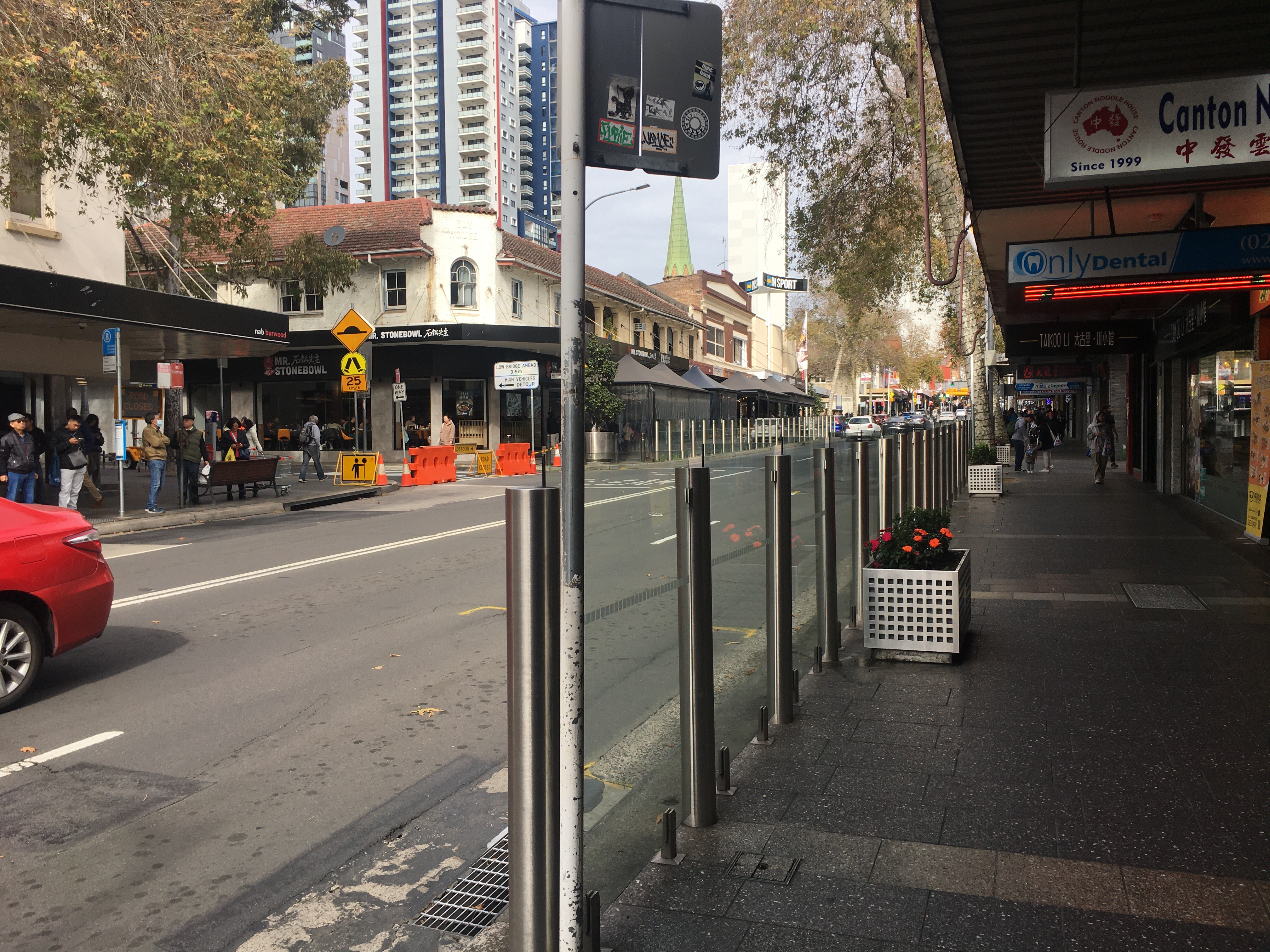 Shot of Burwood Road, looking across and south from a point near the Westfield crossing. Road has 2 lanes each way. The footpaths are wide and lined with shops and restaurants. In the foreground, a long glass pedestrian fence prevents people from crossing the road. On the other side of the road, people are waiting at a bus stop, then after the bus stop, more pedestrian fencing.